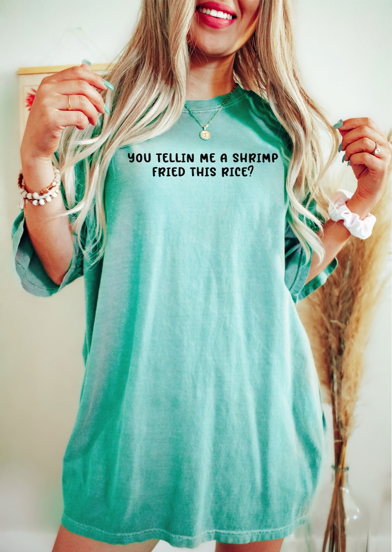 May include: A turquoise t-shirt with the phrase "YOU TELLIN ME A SHRIMP FRIED THIS RICE?" printed in black. The shirt is a relaxed fit and has short sleeves. The image shows the shirt being worn.