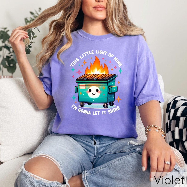 May include: A purple t-shirt with a graphic of a green dumpster on fire with a smiling face and the text "This little light of mine I'm gonna let it shine".