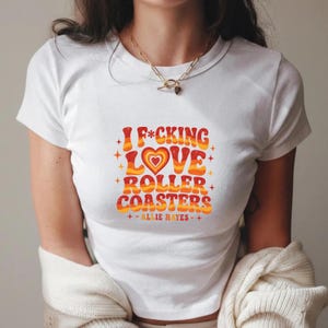 May include: A white short-sleeved t-shirt featuring a graphic print with the text "I F*CKING LOVE ROLLER COASTERS" and "ALLIE HAYES" in a wavy, retro-style font. The design incorporates warm orange, red, and yellow tones with a central heart.