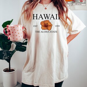 以下が含まれることがあります： 「HAWAII EST 1959 THE ALOHA STATE」という文字と黄色のハイビスカスの花柄がプリントされた白いTシャツです。