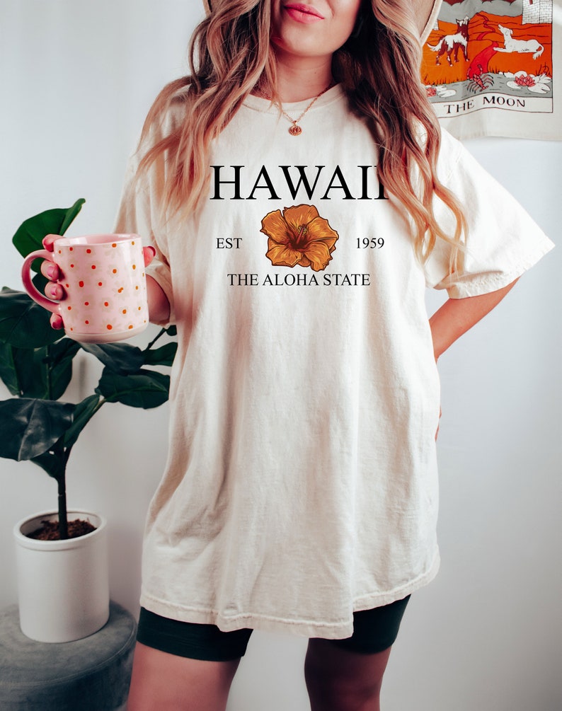 以下が含まれることがあります： 「HAWAII EST 1959 THE ALOHA STATE」という文字と黄色のハイビスカスの花柄がプリントされた白いTシャツです。