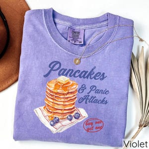 Op de afbeelding: Paars T-shirt met een afbeelding van een stapel pannenkoeken met siroop en boter, en de tekst "Pancakes & Panic Attacks". Het shirt heeft ook de tekst "Buy one get one" in een rode cirkel.