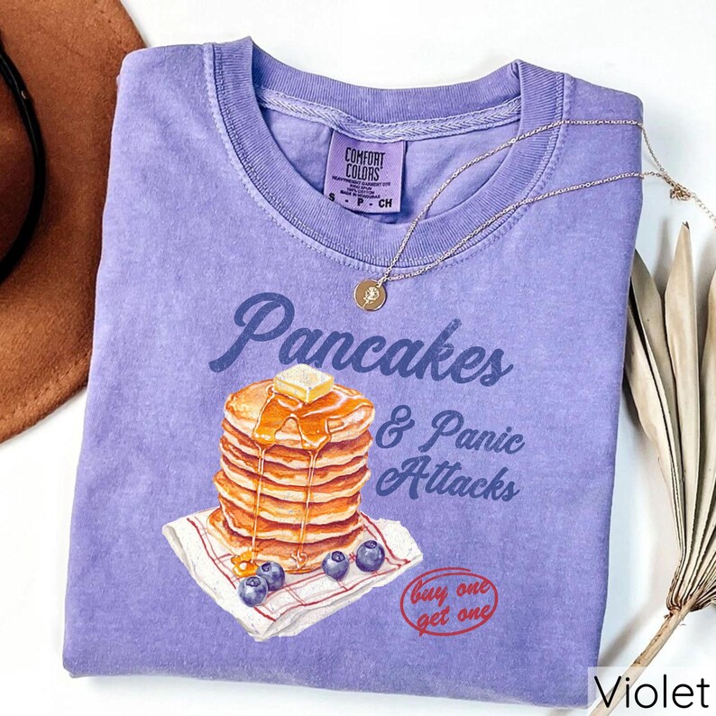 Op de afbeelding: Paars T-shirt met een afbeelding van een stapel pannenkoeken met siroop en boter, en de tekst "Pancakes & Panic Attacks". Het shirt heeft ook de tekst "Buy one get one" in een rode cirkel.