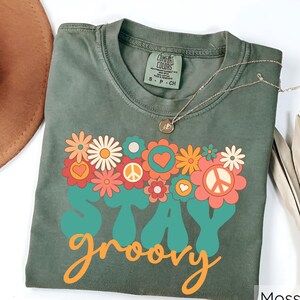 May include: A green t-shirt with a retro-style graphic that says "Stay groovy" with flowers and peace signs. The shirt is made by Comfort Colors.