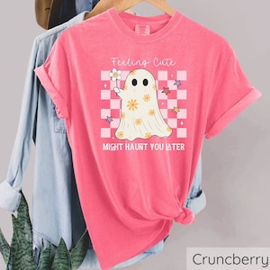 May include: A pink t-shirt with the text "Feeling Cute Might Haunt You Later" and a cartoon ghost holding a flower. The ghost is on a checkered background with stars. The shirt is hanging on a wooden rack.