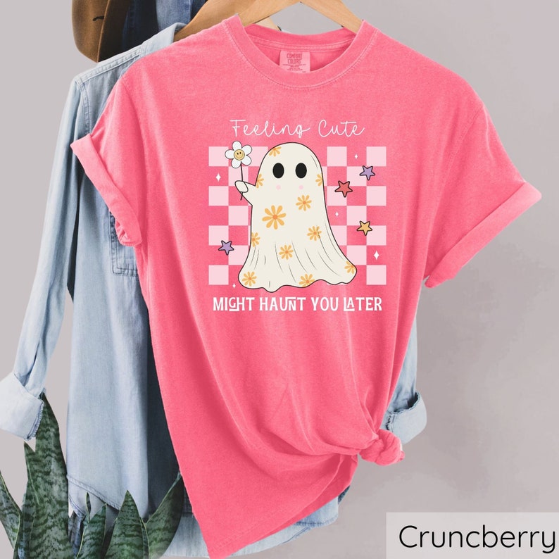 May include: A pink t-shirt with the text "Feeling Cute Might Haunt You Later" and a cartoon ghost holding a flower. The ghost is on a checkered background with stars. The shirt is hanging on a wooden rack.