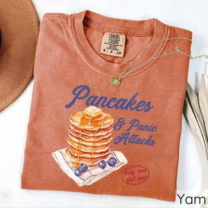 Op de afbeelding: Een roestkleurig T-shirt met een afbeelding van een stapel pannenkoeken met siroop en boter, en de tekst "Pancakes & Panic Attacks". Het shirt heeft ook de tekst "Buy one get one" in een cirkel.