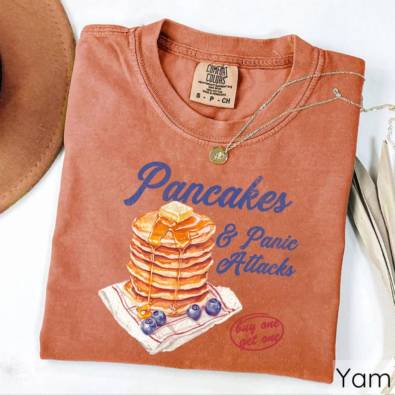 Op de afbeelding: Een roestkleurig T-shirt met een afbeelding van een stapel pannenkoeken met siroop en boter, en de tekst "Pancakes & Panic Attacks". Het shirt heeft ook de tekst "Buy one get one" in een cirkel.