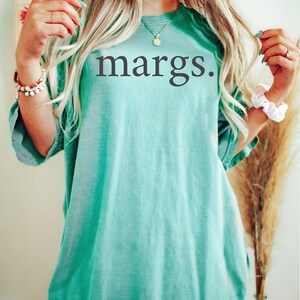 Puede incluir: Una camiseta de gran tama&ntilde;o de color azul turquesa con la palabra "margs." impresa en negro.