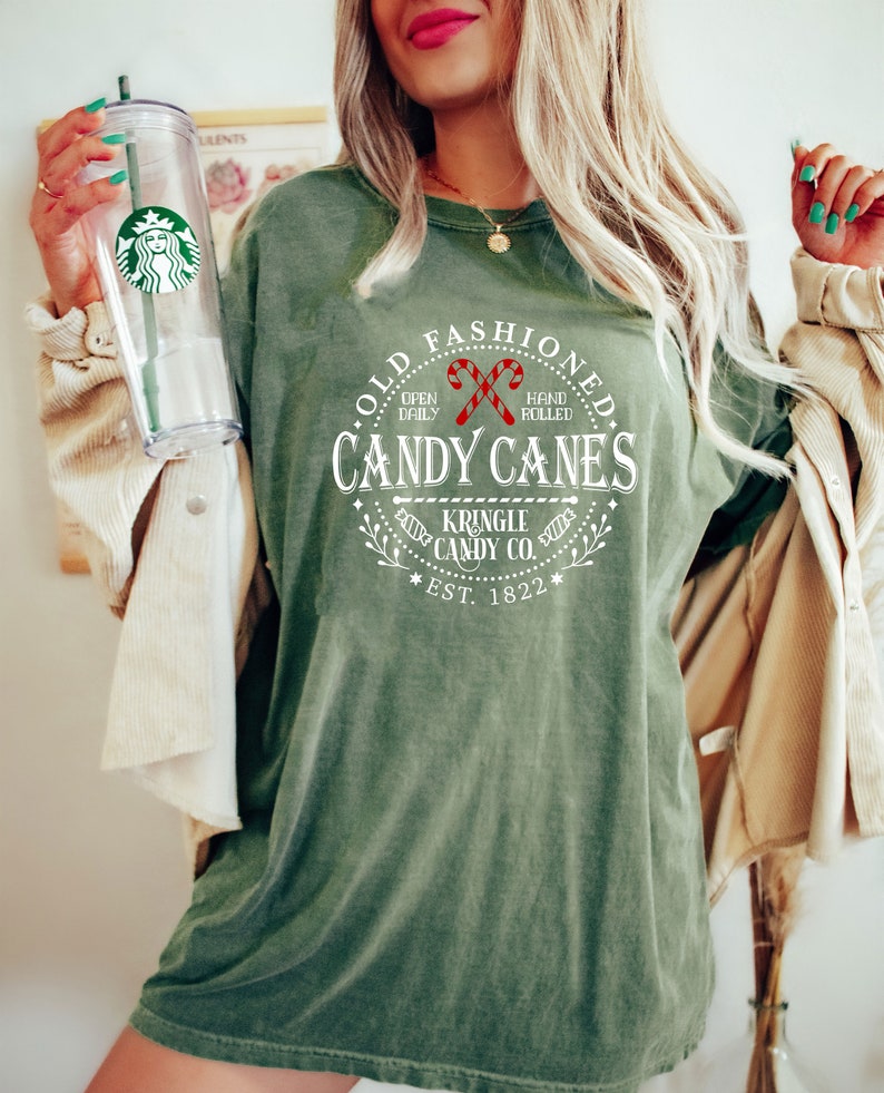 Pu&ograve; includere: T-shirt verde oliva con la scritta "OLD FASHIONED CANDY CANES" in un design circolare, con grafica di bastoncini di zucchero. La maglietta mostra anche "KRINGLE CANDY CO. EST. 1822". Nella foto &egrave; presente un bicchiere trasparente Starbucks con una cannuccia verde.