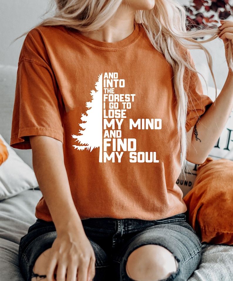 K&ouml;nnte beinhalten: Ein orangefarbenes T-Shirt mit einer wei&szlig;en Silhouette eines Kiefernbaums und dem Text "AND INTO THE FOREST I GO TO LOSE MY MIND AND FIND MY SOUL".