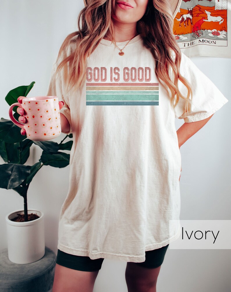 Puede incluir: Una mujer lleva una camiseta de color crema con el texto "God is Good" en una fuente de estilo vintage con un dise&ntilde;o de rayas arco&iacute;ris. La camiseta es de gran tama&ntilde;o y la mujer lleva pantalones cortos negros. El fondo es una pared blanca.