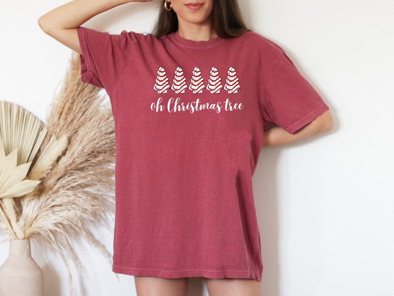 May include: A maroon t-shirt with the phrase "oh Christmas tree" in white script. Above the text are four white Christmas tree designs with red accents. The shirt has short sleeves and a relaxed fit.