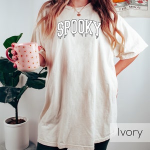 May include: Ivory-colored t-shirt with the word "SPOOKY" in a black, dripping font. The shirt is a relaxed fit. The model is wearing dark green shorts and holding a pink mug with orange polka dots. A plant is in the background.