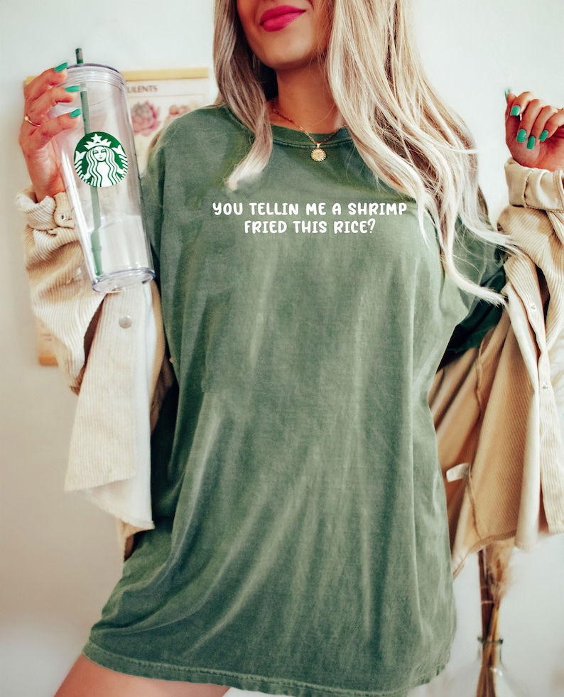 May include: Olive green t-shirt with the text "YOU TELLIN ME A SHRIMP FRIED THIS RICE?" in white. A clear Starbucks tumbler with a green straw and logo is held in the left hand. The person is wearing a tan jacket.