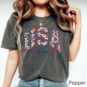 May include: Dark grey t-shirt with the word "USA" in a floral pattern of red, white, and blue. The text "EST 1776" is below the word "USA". The shirt has short sleeves.