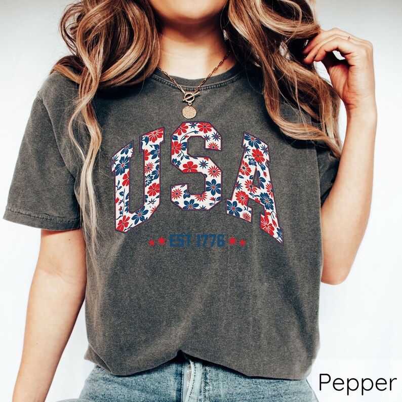 May include: Dark grey t-shirt with the word "USA" in a floral pattern of red, white, and blue. The text "EST 1776" is below the word "USA". The shirt has short sleeves.
