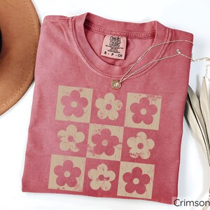 May include: A crimson red t-shirt with a vintage-style floral pattern in a tan colour. The shirt has a distressed look and features the text "COMFORT COLORS" on the tag.