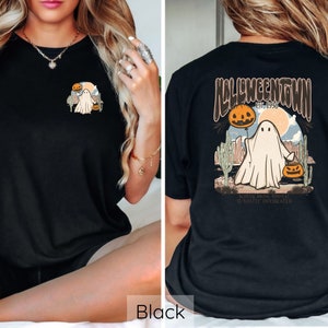 May include: Black t-shirt featuring a Halloween-themed graphic. The front displays a small design of a ghost, pumpkin, and desert landscape. The back showcases a larger design with the text "Halloweentown Est. 1998" and a ghost holding a pumpkin balloon.