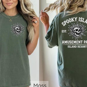 May include: Moss green t-shirt with a skull graphic on the front and back. The front features a skull within a starburst design. The back of the shirt displays the text "Spooky Island Amusement Park Island Resort" with a skull graphic.