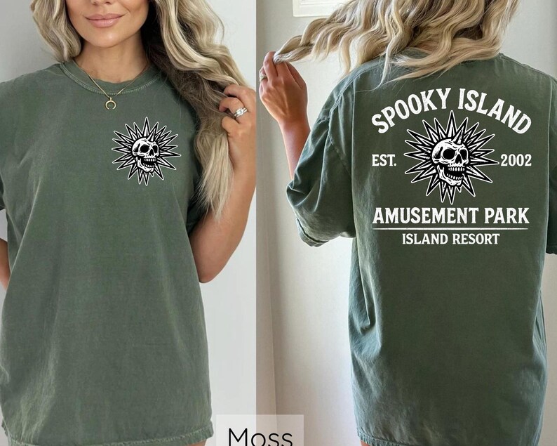 May include: Moss green t-shirt with a skull graphic on the front and back. The front features a skull within a starburst design. The back of the shirt displays the text "Spooky Island Amusement Park Island Resort" with a skull graphic.