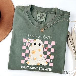 May include: Moss green t-shirt with a ghost graphic holding a flower. The ghost has orange flower details and is set against a pink and white checkered background. Text reads "Feeling Cute Might Haunt You Later". A gold necklace is draped on the shirt.