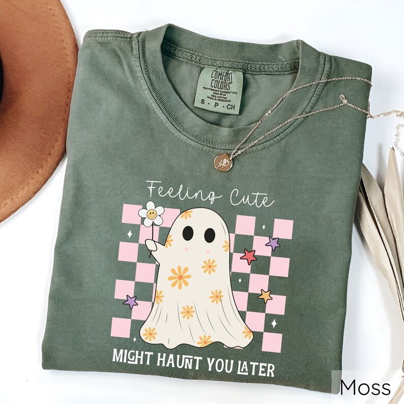 May include: Moss green t-shirt with a ghost graphic holding a flower. The ghost has orange flower details and is set against a pink and white checkered background. Text reads "Feeling Cute Might Haunt You Later". A gold necklace is draped on the shirt.