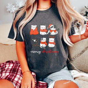 May include: A charcoal gray t-shirt featuring a graphic of six cartoon cats in winter attire, including scarves, a Santa hat, and reindeer antlers. One cat is wrapped in a red blanket, and another is tangled in string lights. The text reads meowy christmas.
