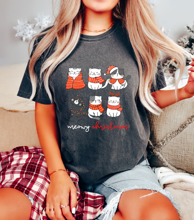 May include: A charcoal gray t-shirt featuring a graphic of six cartoon cats in winter attire, including scarves, a Santa hat, and reindeer antlers. One cat is wrapped in a red blanket, and another is tangled in string lights. The text reads meowy christmas.