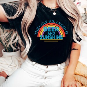 May include: Black t-shirt with a rainbow graphic and the text "THE ONLY B.S. I NEED IS BEER AND SUNSHINE". The shirt is paired with white pants. A gold necklace and rings are visible.