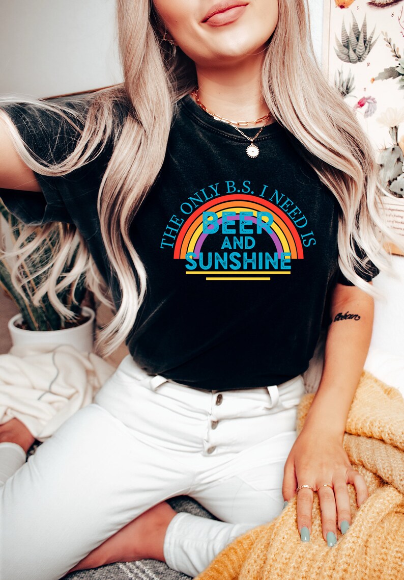 May include: Black t-shirt with a rainbow graphic and the text "THE ONLY B.S. I NEED IS BEER AND SUNSHINE". The shirt is paired with white pants. A gold necklace and rings are visible.