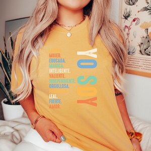May include: A yellow t-shirt with the text "YO SOY" in colorful letters. The text is surrounded by a list of Spanish words describing a woman, including "MUJER", "EDUCADA", "MAGICA", "INTELIGENTE", "VALIENTE", "INDEPENDIENTE", "ORGULLOSA", "LEAL", "FUERTE", and "AMOR".