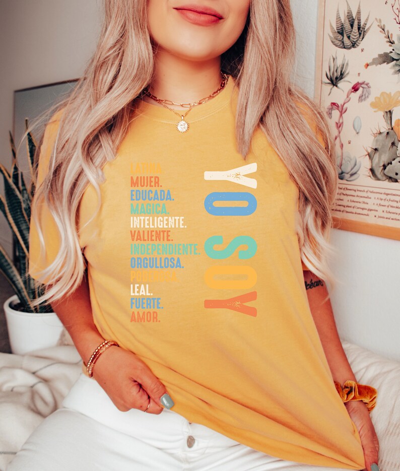 May include: A yellow t-shirt with the text "YO SOY" in colorful letters. The text is surrounded by a list of Spanish words describing a woman, including "MUJER", "EDUCADA", "MAGICA", "INTELIGENTE", "VALIENTE", "INDEPENDIENTE", "ORGULLOSA", "LEAL", "FUERTE", and "AMOR".