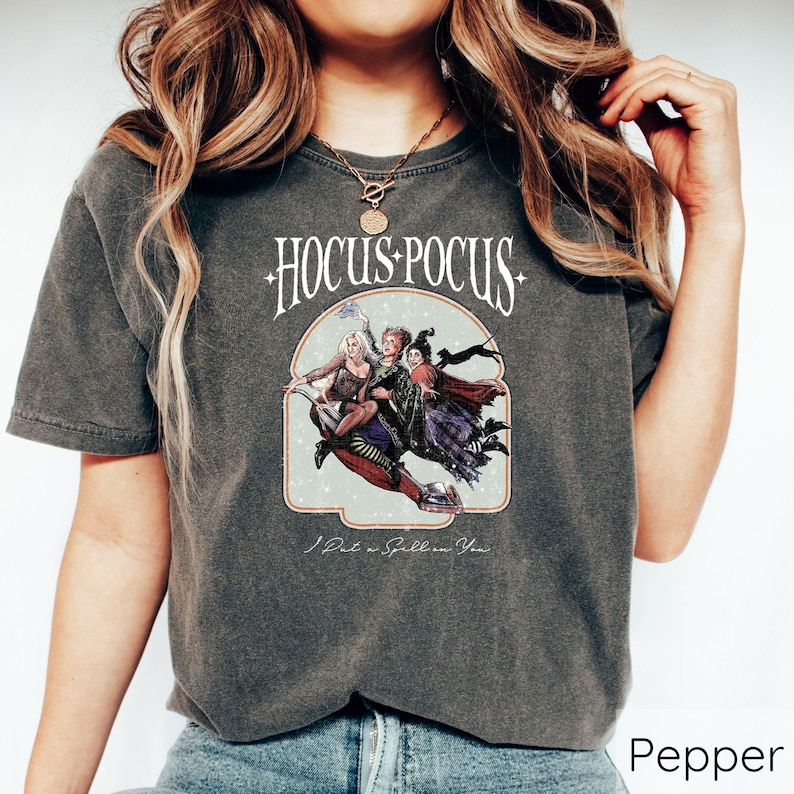 Puede incluir: Camiseta color pimienta con el texto "Hocus Pocus" en una fuente estilizada sobre una ilustraci&oacute;n de tres brujas en una escoba. El gr&aacute;fico incluye la frase "I Put a Spell on You."