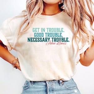 May include: Cream-colored t-shirt with the text "GET IN TROUBLE. GOOD TROUBLE. NECESSARY TROUBLE. John Lewis" in teal and red lettering. The shirt is made of a soft material and is being worn by a person.