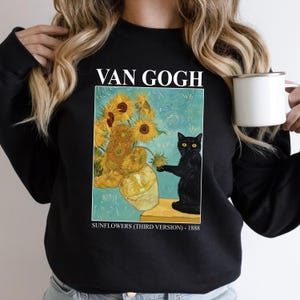 May include: Black sweatshirt featuring a print of Van Gogh's "Sunflowers" painting with a black cat. The text "VAN GOGH" is above the image, and "SUNFLOWERS (THIRD VERSION) - 1888" is below. A white mug is held in the right hand.