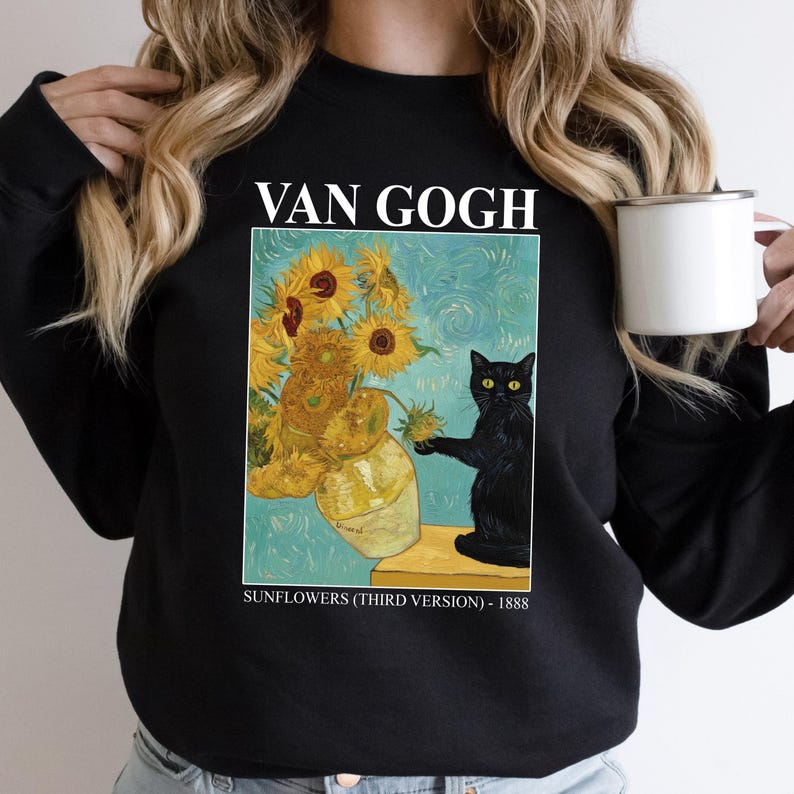 May include: Black sweatshirt featuring a print of Van Gogh's "Sunflowers" painting with a black cat. The text "VAN GOGH" is above the image, and "SUNFLOWERS (THIRD VERSION) - 1888" is below. A white mug is held in the right hand.