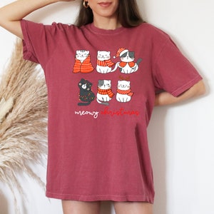 May include: A dark red short-sleeve t-shirt featuring a graphic of six cartoon cats wearing winter accessories like scarves, a Santa hat, and reindeer antlers, with the text "meowy christmas" printed in white and red script below the cats.