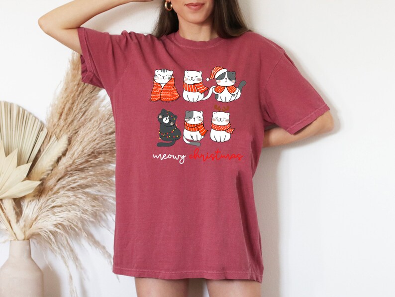 May include: A dark red short-sleeve t-shirt featuring a graphic of six cartoon cats wearing winter accessories like scarves, a Santa hat, and reindeer antlers, with the text "meowy christmas" printed in white and red script below the cats.
