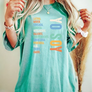 May include: A turquoise blue t-shirt with the text "YO SOY" in rainbow colors. The text is surrounded by a list of Spanish words describing a woman's qualities, including "Latina", "Mujer", "Educada", "Magica", "Inteligente", "Valiente", "Independiente", "Orgullosa", "Poderosa", "Leal", "Fuerte", and "Amor."
