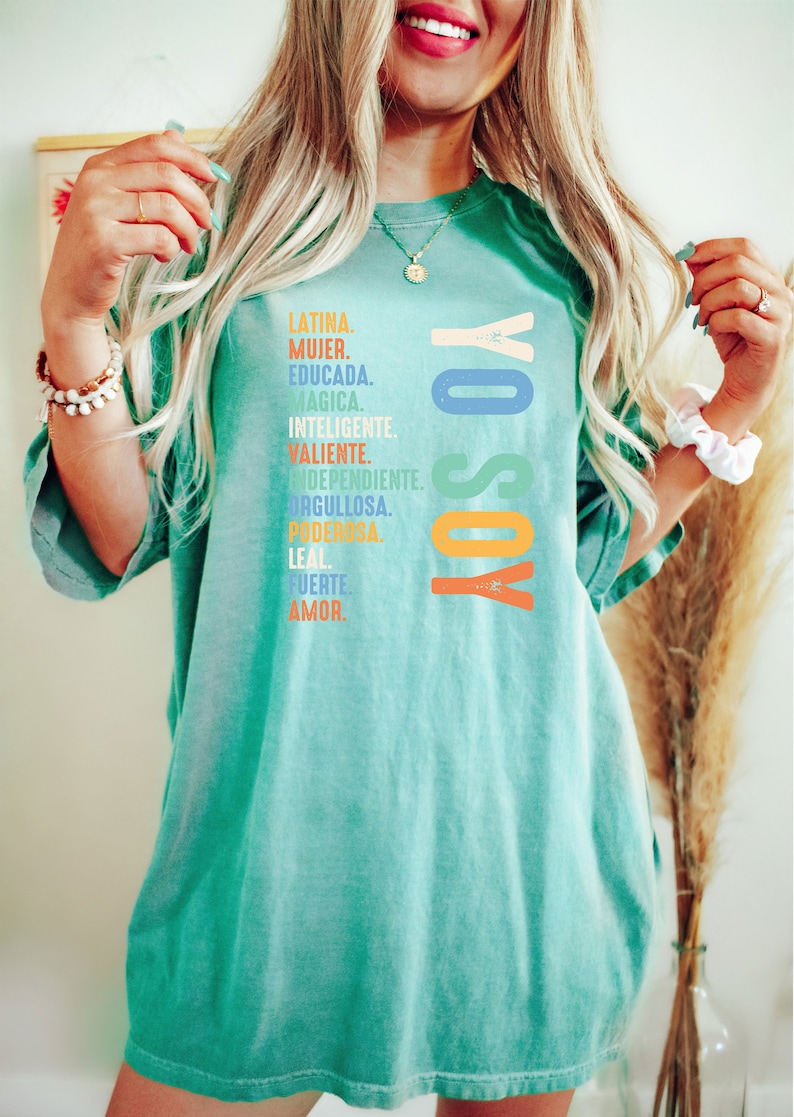 May include: A turquoise blue t-shirt with the text "YO SOY" in rainbow colors. The text is surrounded by a list of Spanish words describing a woman's qualities, including "Latina", "Mujer", "Educada", "Magica", "Inteligente", "Valiente", "Independiente", "Orgullosa", "Poderosa", "Leal", "Fuerte", and "Amor."