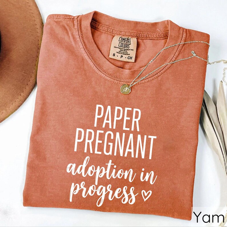 Pode incluir: Uma camiseta cor de terracota com as palavras "PAPER PREGNANT adoption in progress" em escrita branca. A camiseta tem gola redonda e mangas curtas. Um colar de ouro com um pingente est&aacute; sobre a camiseta.