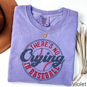 May include: A purple t-shirt with a distressed look featuring a white baseball with red stitching inside a red circle with the text "There's No Crying In Baseball".