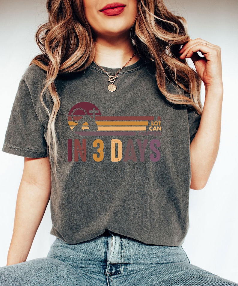 Może przedstawiać: Szary t-shirt z retro grafiką przedstawiającą krzyż, zach&oacute;d słońca i napis "A LOT CAN HAPPEN IN 3 DAYS".