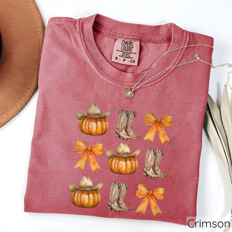 May include: Crimson-colored t-shirt with a crew neck. The shirt features a repeating pattern of pumpkins with cowboy hats, cowboy boots, and orange bows. A silver necklace with a pendant is draped across the shirt.