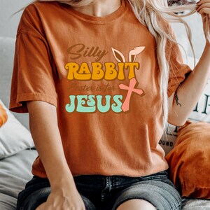 May include: A woman wearing a burnt orange t-shirt with the text "Silly Rabbit Easter is for Jesus" in a retro font. The text is in shades of orange, yellow, pink, and blue.