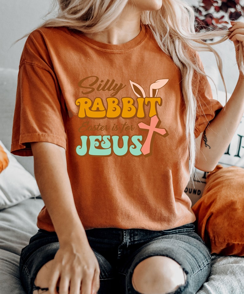 May include: A woman wearing a burnt orange t-shirt with the text "Silly Rabbit Easter is for Jesus" in a retro font. The text is in shades of orange, yellow, pink, and blue.