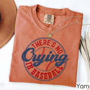 May include: A burnt orange t-shirt with a distressed, vintage look. The shirt features a blue and red graphic with a baseball and the text "There's No Crying In Baseball".