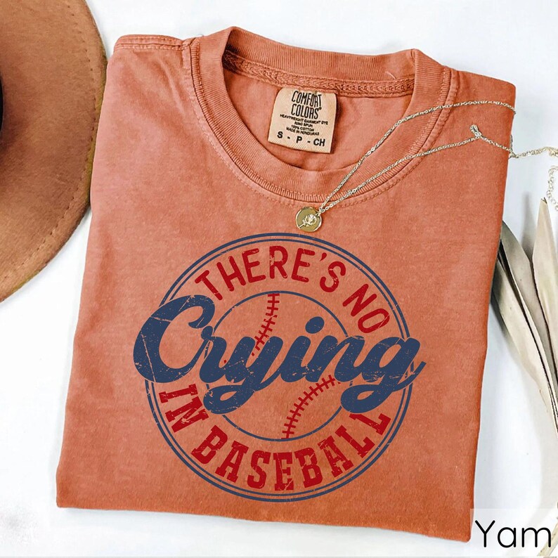 May include: A burnt orange t-shirt with a distressed, vintage look. The shirt features a blue and red graphic with a baseball and the text "There's No Crying In Baseball".