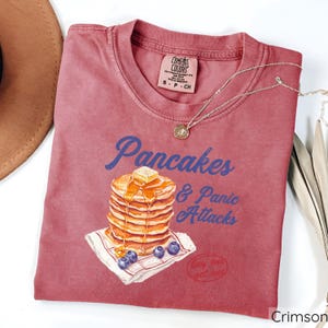 Op de afbeelding: Crimsonrode T-shirt met een afbeelding van een stapel pannenkoeken met siroop en boter, en de tekst "Pancakes & Panic Attacks".